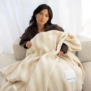 A person sits on a light sofa, holding The Wool Stripe Throw - Pale Taupe with cream and beige stripes and fringe edges. A white label is visible on the blanket, and white curtains hang in the background. Gate21.ee