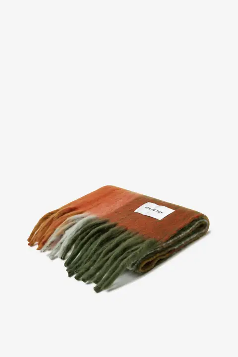 The Stockholm Scarf - Mossy Spring is a folded wool scarf with long fringe and a gradient from rust orange to green. A white Arctic Fox Stockholm label is visible on the plain white background. Gate21.ee