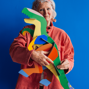 An older woman in a pink corduroy jacket and patterned headscarf stands against a blue background, holding the colourful Deluxe T-Rex cardboard model by StudioROOF in her arms. Gate21.ee