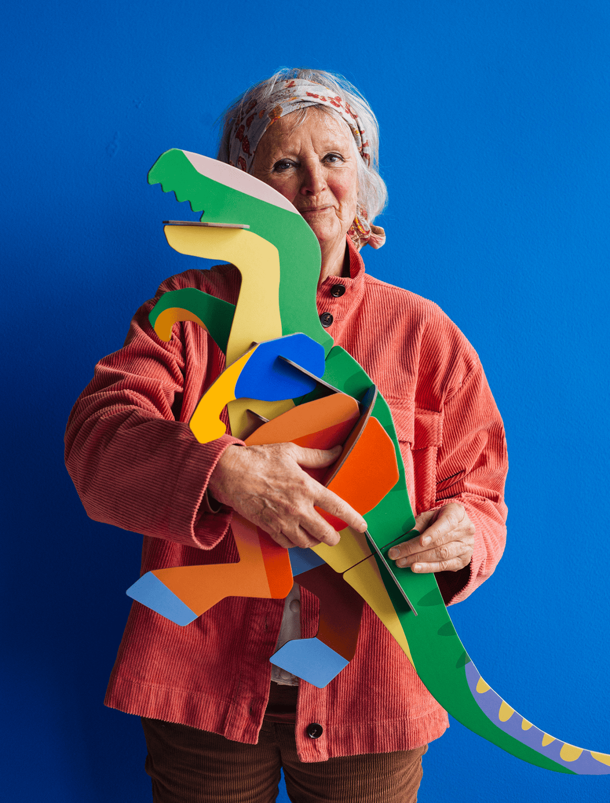An older woman in a pink corduroy jacket and patterned headscarf stands against a blue background, holding the colourful Deluxe T-Rex cardboard model by StudioROOF in her arms. Gate21.ee