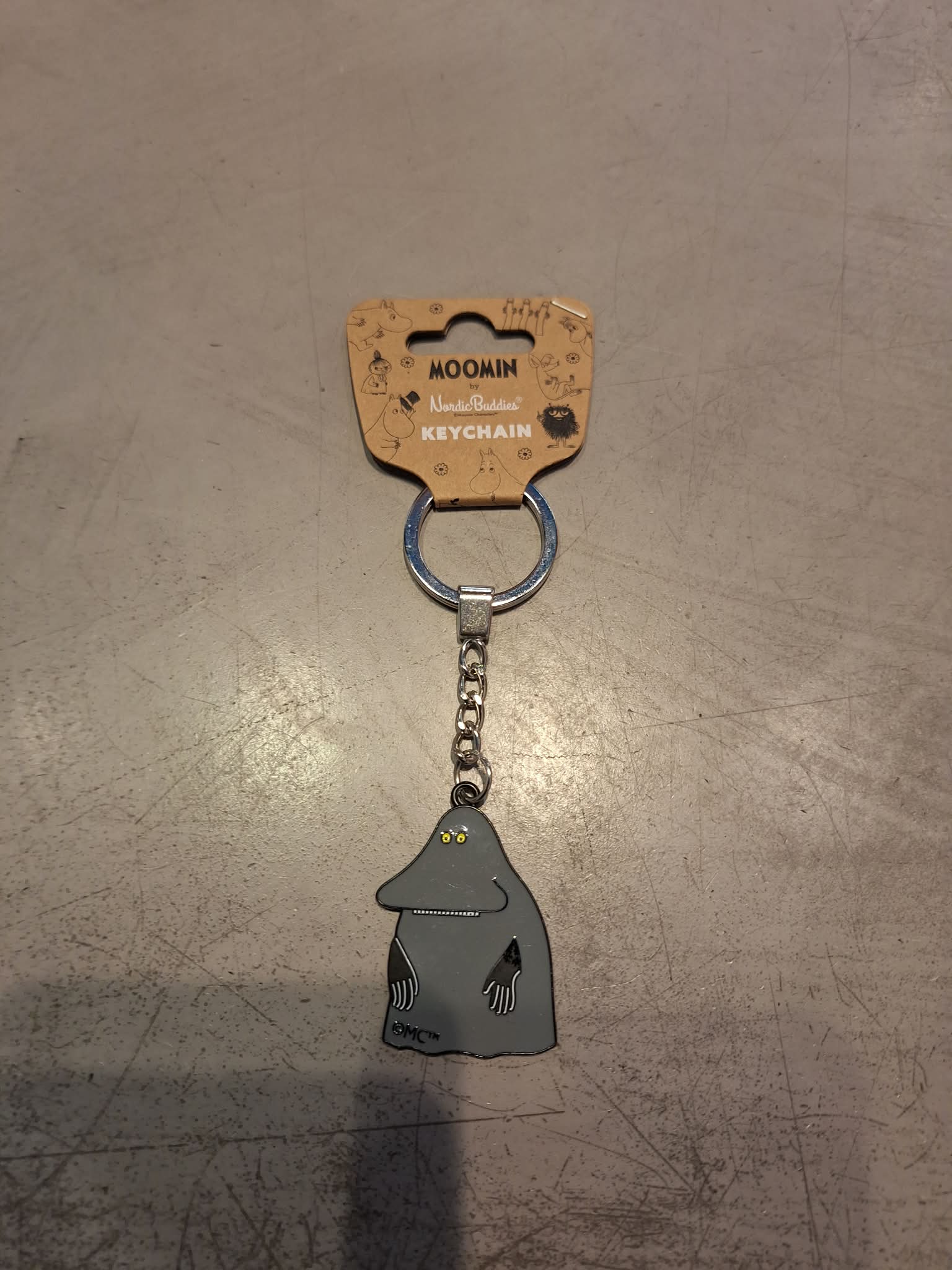 A Moomin Keyring - Gnork featuring a grey Gnork figure attached to a silver chain and ring, displayed on a grey surface. Ideal for collectors of Moomin keyrings, the packaging reads: MOOMIN Nordicbuddies KEYRING. Gate21.ee