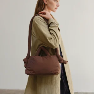 A person wearing a long beige coat holds the Mini Ona Soft Bag - Chocolate, a brown padded handbag, over their shoulder against a plain light background. Gate21.ee