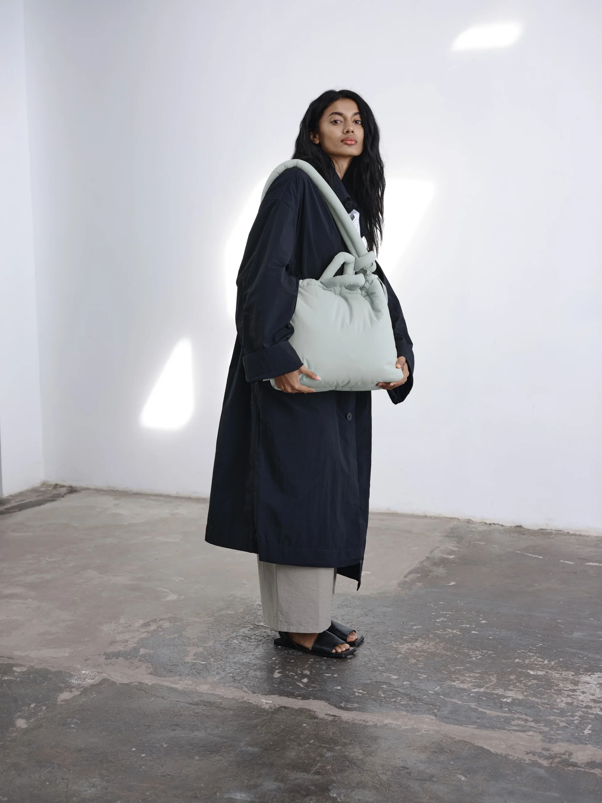 A person with long dark hair stands indoors on a concrete floor, wearing a dark coat, light trousers, and black sandals, carrying an Ona Soft Bag - Pearl Grey over one shoulder against a plain white wall. Gate21.ee