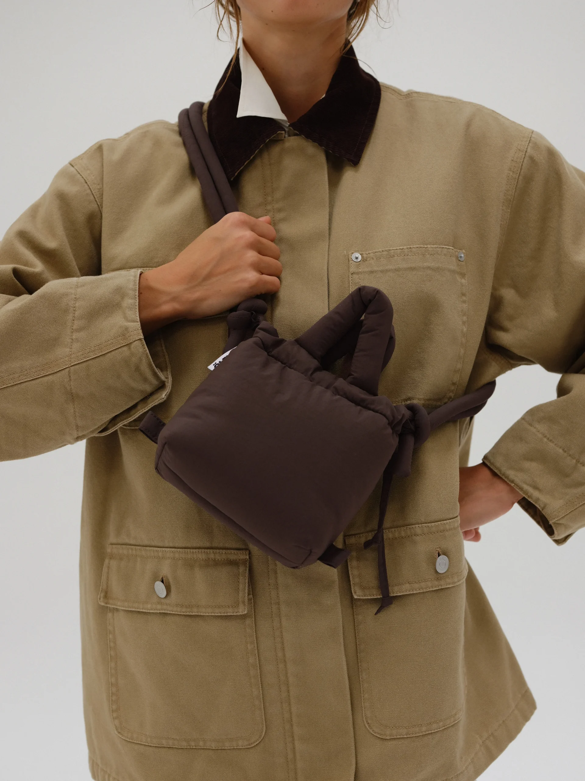 A person in a tan utility jacket with dark brown accents holds the Micro Ona Soft Bag - Chocolate, a small, padded dark brown shoulder bag, across their chest against a plain light background. Gate21.ee