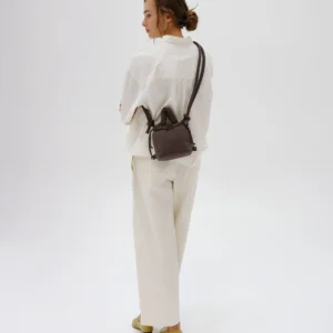 A person stands on a white background, wearing a loose white shirt, wide white trousers, and light shoes. They have the Micro Ona Soft Bag - Chocolate slung over their shoulder and look to the side with arms crossed. Gate21.ee