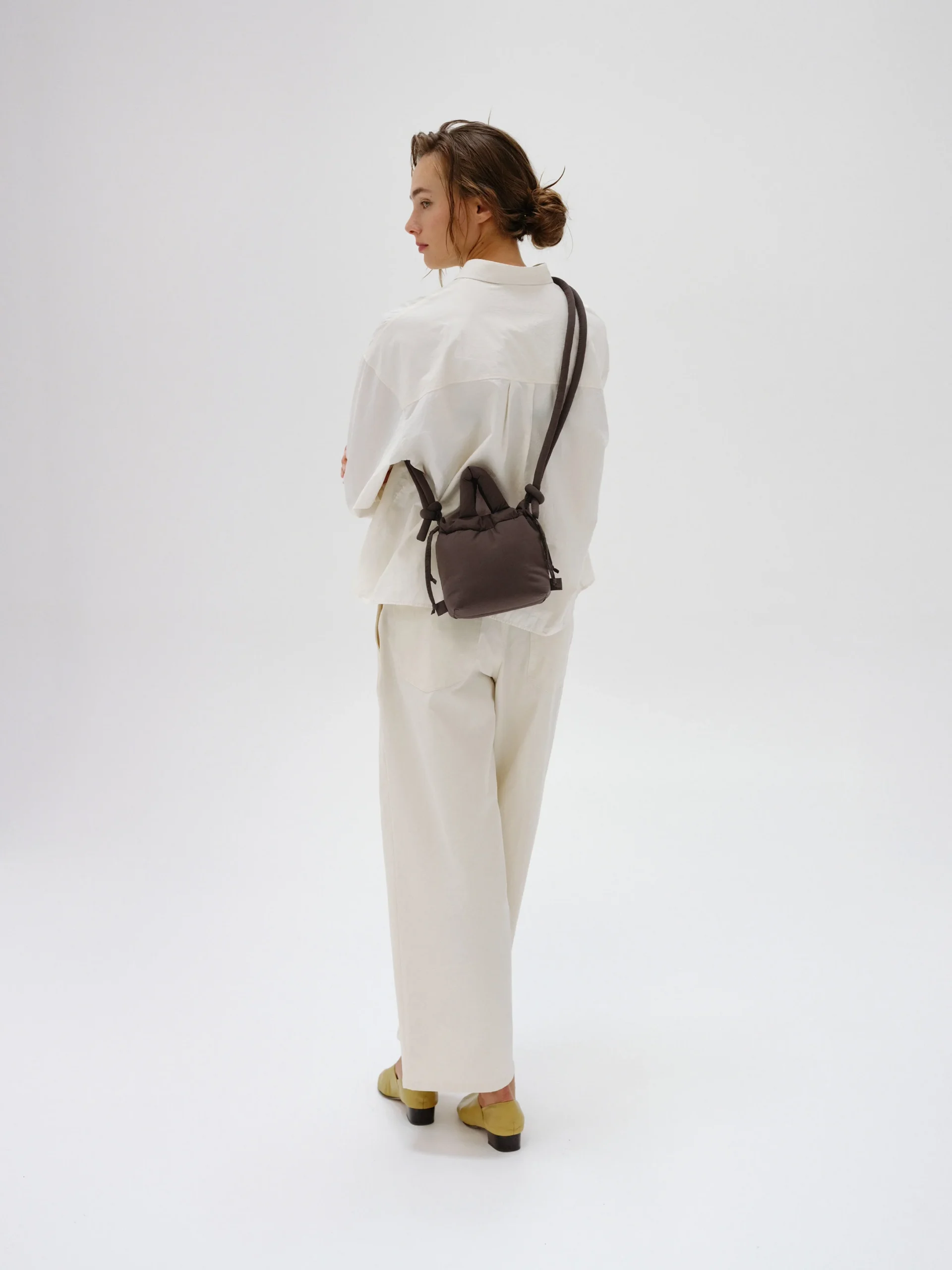 A person stands on a white background, wearing a loose white shirt, wide white trousers, and light shoes. They have the Micro Ona Soft Bag - Chocolate slung over their shoulder and look to the side with arms crossed. Gate21.ee