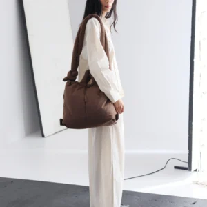A woman with long dark hair, dressed in a loose white outfit and black shoes, stands indoors holding the Ona Soft Bag - Chocolate by ÖLEND. A tilted mirror and light fitting appear in the background. Gate21.ee