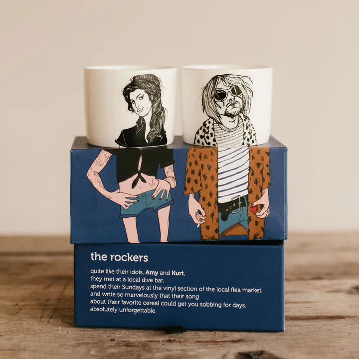 The Rockers Espresso set features two white cups with illustrated portraits of a man and a woman, displayed on a blue box with matching artwork and narrative text, all shown on a wooden surface. Gate21.ee