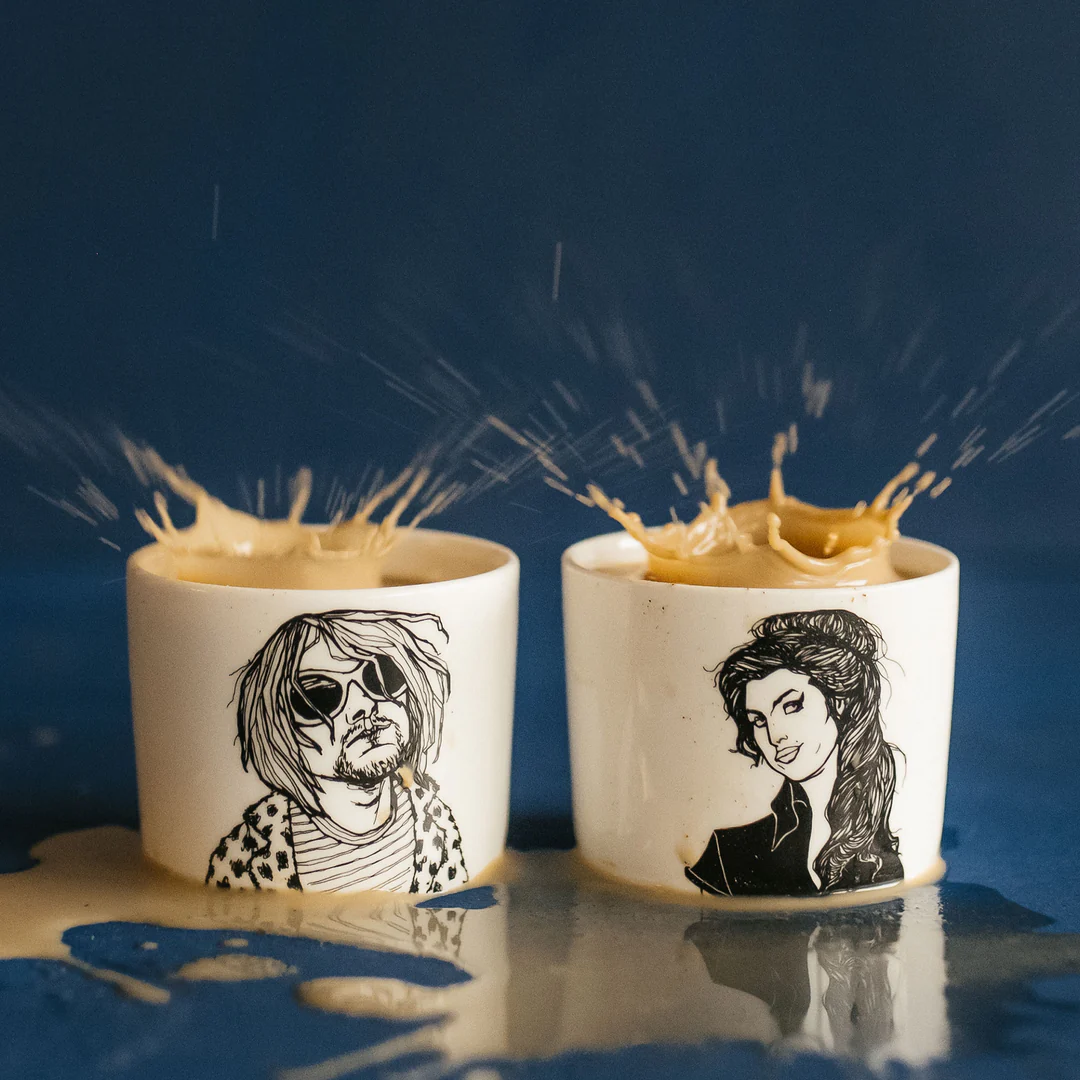 The Rockers Espresso set features two white mugs with illustrated portraits, coffee splashing out and pooling at their base against a vibrant blue background. Gate21.ee