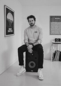 A man with short hair and a beard sits on a large speaker in a minimalist room. He wears a sweatshirt, dark trousers, and white trainers. Behind him is the (Sittin’ on) The Dock of the Bay - Lyrics Poster among framed art and records. Gate21.ee