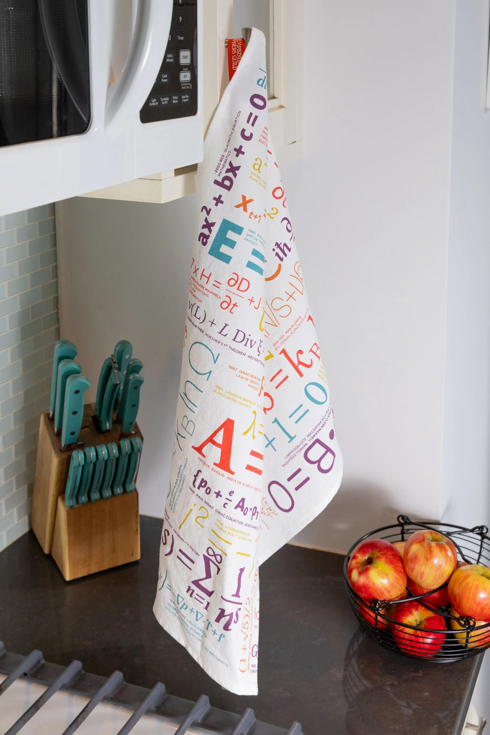 The Equations - Tea Towel from the Unemployed Philosophers Guild, featuring colourful maths equations and symbols, hangs from a cupboard handle by the microwave above a knife block, wire fruit basket with apples, and hob. Gate21.ee