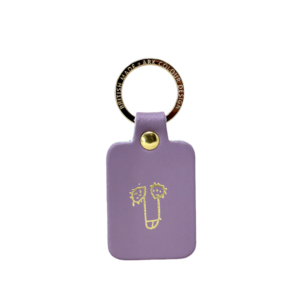 The Dick Key Ring - Lilac is a rectangular lavender keyring with a gold-toned ring engraved with “BRITISH HANDMADE DARK COLOUR DESIGN” and a simple, doodle-style face in gold on the front, inspired by Estonian design. Gate21.ee