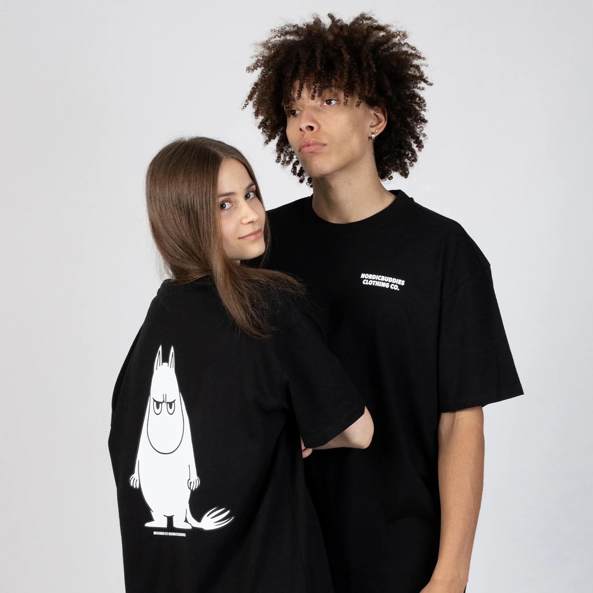 A young woman and man wear black "Moomin Ts - Moomintroll Temper" shirts. Hers shows a large white Moomintroll on the back; his has small white text on the front. Both face the camera against a plain background. Gate21.ee