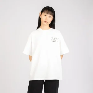 A person with straight black hair and a fringe stands against a plain light background, wearing a loose white Moomin Ts - Novels T-shirt with a small chest graphic and black trousers. Gate21.ee