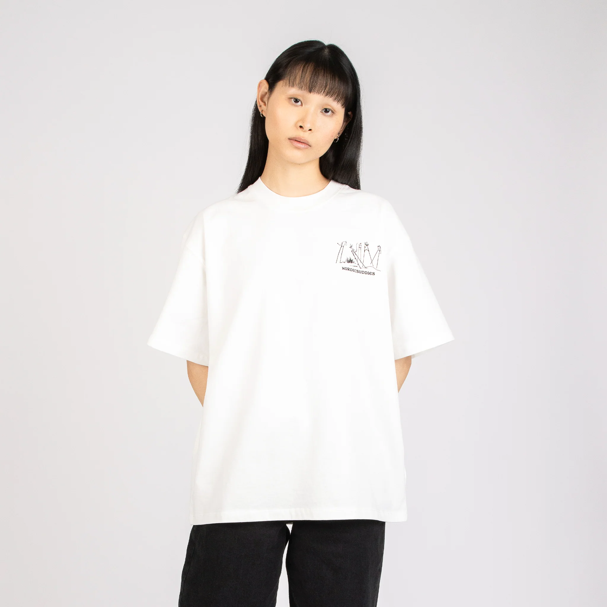 A person with straight black hair and a fringe stands against a plain light background, wearing a loose white Moomin Ts - Novels T-shirt with a small chest graphic and black trousers. Gate21.ee