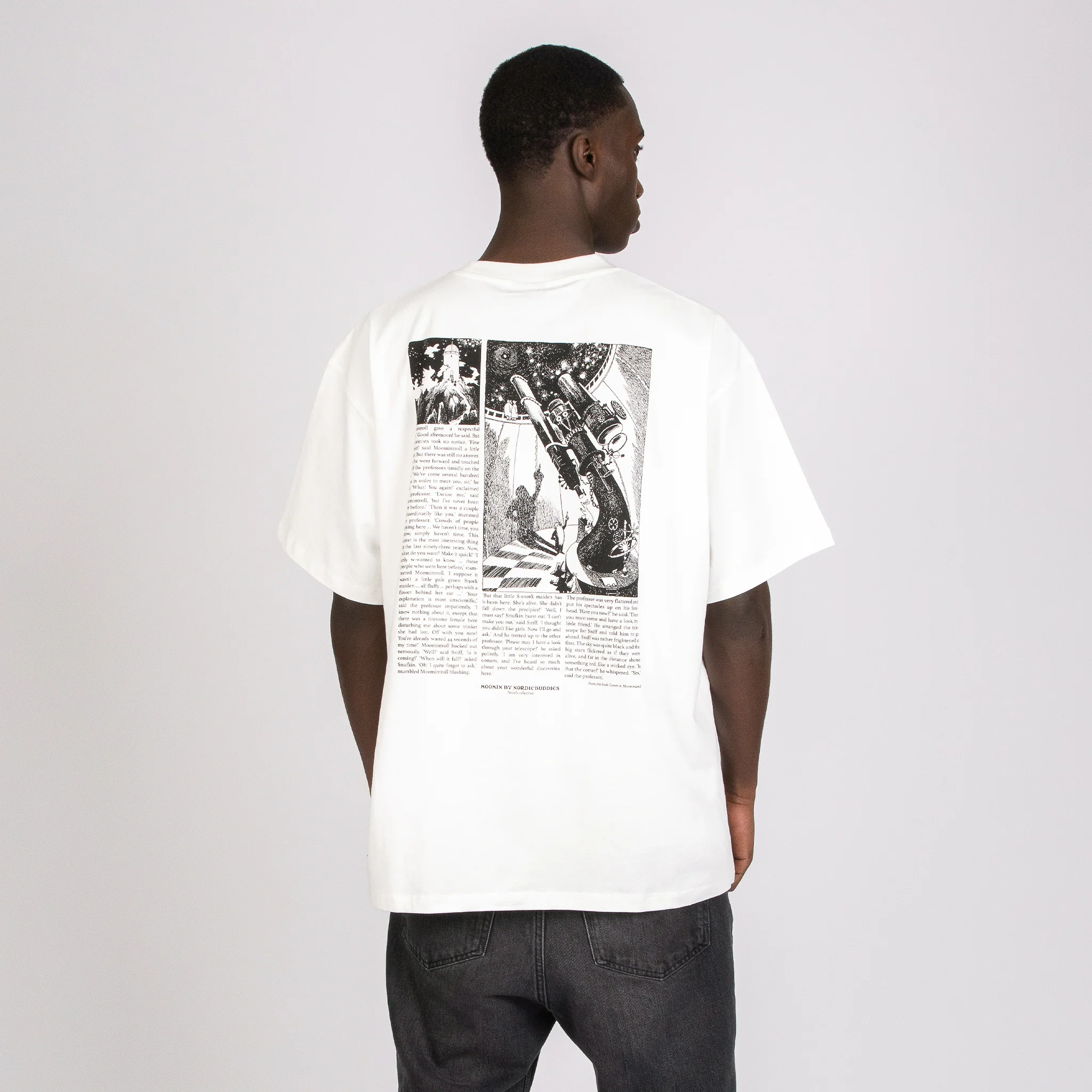 A person wearing dark jeans faces away, showcasing the Moomin Ts - Novels white T-shirt with a black-and-white graphic and text on the back against a plain light grey background. Gate21.ee