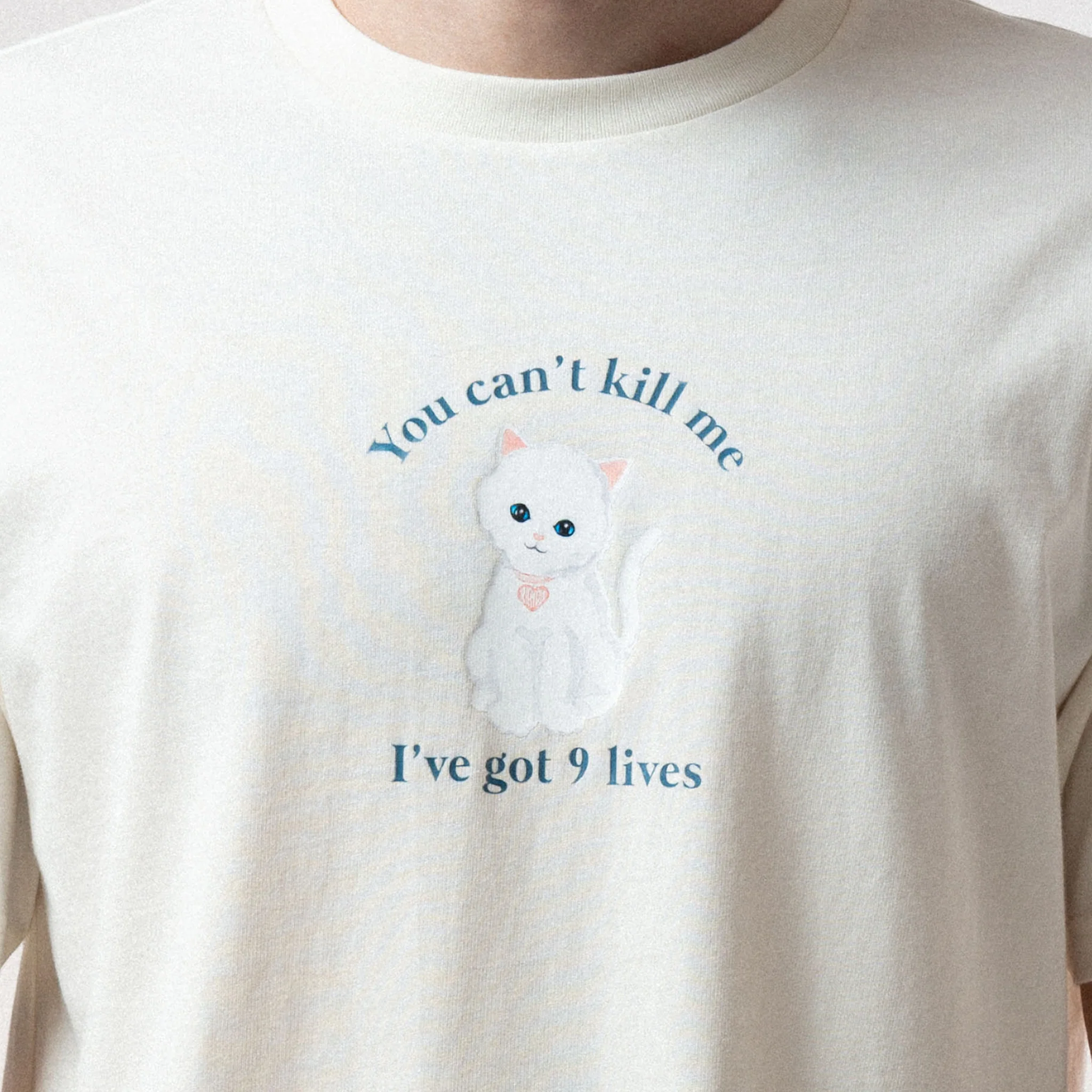 A person wearing the 9 Lives Tee - Bone, a cream-coloured shirt featuring a white cat illustration and the phrase "You can't kill me I've got 9 lives" on the front, inspired by iconic RipnDip style. Gate21.ee