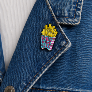 A close-up of a denim jacket features the “Fries before Guys - Pin,” a colourful enamel pin shaped like a box of chips with “FRIES BEFORE GUYS” text. A white shirt is visible under the jacket. Gate21.ee