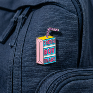 The Fresh Boy Tears - Pin, a colourful enamel juice box with a straw and "FRESH BOY TEARS" in pink, yellow, and blue letters, is attached to a dark blue fabric rucksack. Gate21.ee