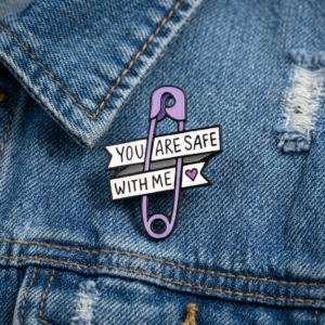The Safe with Me - Pin, a purple safety pin enamel pin, is fastened to blue denim and features two white banners with black text saying “YOU ARE SAFE WITH ME” and a small purple heart on the right side. Gate21.ee