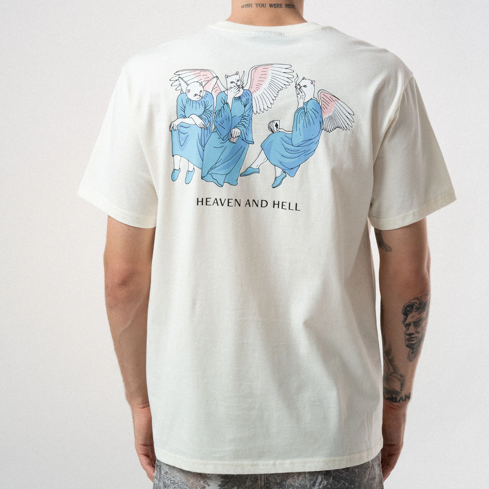 A person wearing a white Heaven and Hell Tee - Bone, featuring three winged, robed animal figures with “HEAVEN AND HELL” text beneath. Tattoos are visible on their arms and neck. Gate21.ee