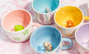 Five pastel-coloured ceramic mugs sit on a pink marbled surface. Each mug has a different animal figurine inside, including a frog, dog, elephant, bear, and a cow or bull. Gate21.ee