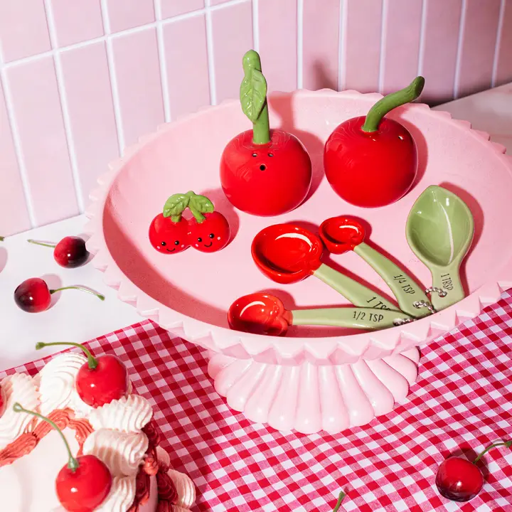 A pink cake stand holds Cherry - Salt & Pepper Shakers, a small cherry dish with a face, and measuring spoons. A slice of cake and loose cherries rest on a pink gingham cloth in front of a pink tiled wall. Gate21.ee