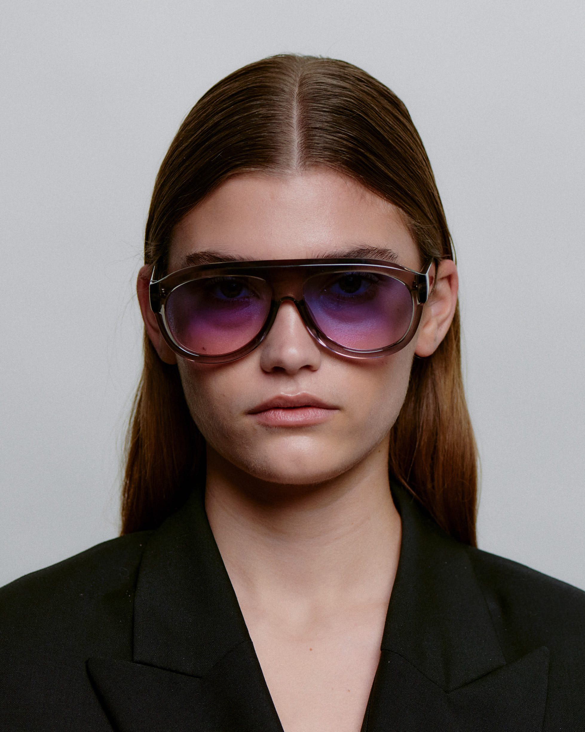A person with straight brown hair wears large Rio - Grey transparent A. Kjærbede sunglasses and a black blazer, facing the camera against a plain light background. Gate21.ee