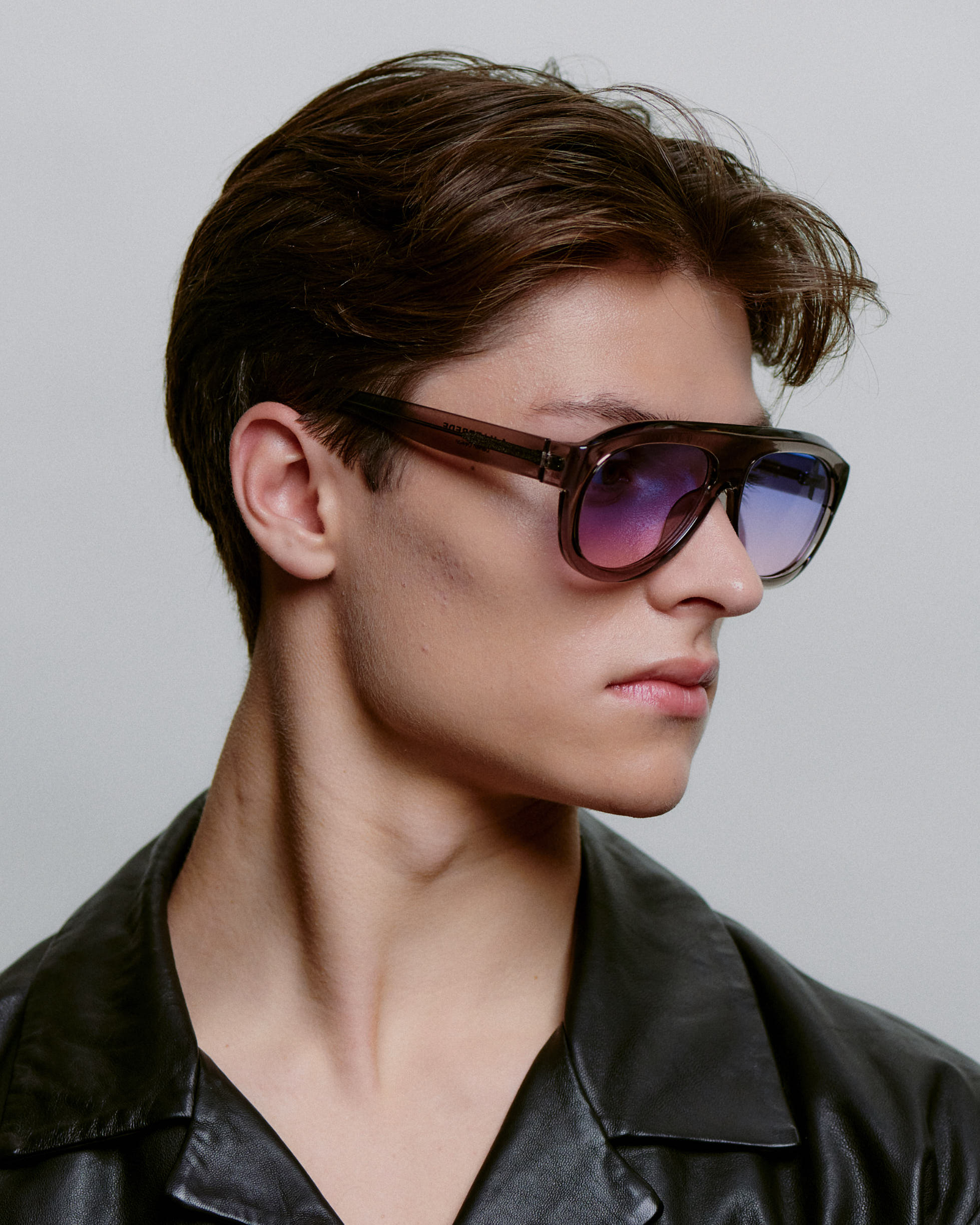 A person with short brown hair wearing a black leather jacket and large Rio - Grey transparent sunglasses by A. Kjærbede looks to the side against a plain light background. Gate21.ee