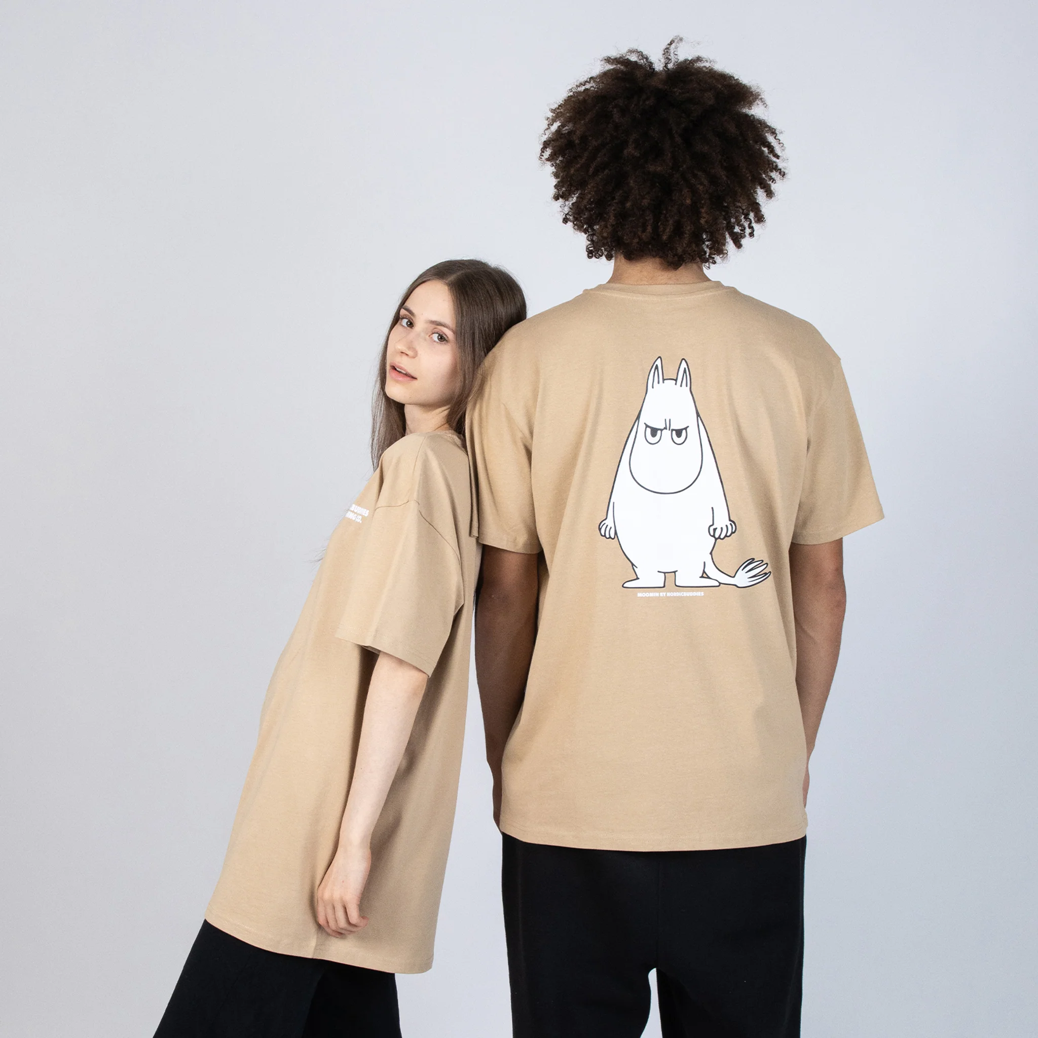 Two people stand back to back in beige "Moomin Ts – Moomintroll Temper" shirts. The left person faces forwards; the right shows off a white Moomintroll cartoon printed on the shirt's back. Gate21.ee