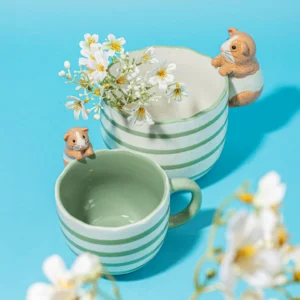 Two green-and-white striped ceramic "Guinea Pig - Cup" cups, one featuring painted hamster figures on the rim, are shown on a blue background with white flowers placed in and around them. Gate21.ee