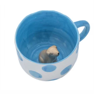 A white ceramic mug with large blue polka dots, a blue handle, and a small ceramic dog figurine fixed at the bottom inside. Product name: Surprise Dog - Cup. Gate21.ee