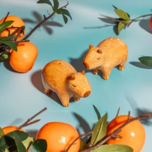 Two Capybara Salt & Pepper Shakers sit on a light blue surface, surrounded by artificial oranges and green leaves. Gate21.ee