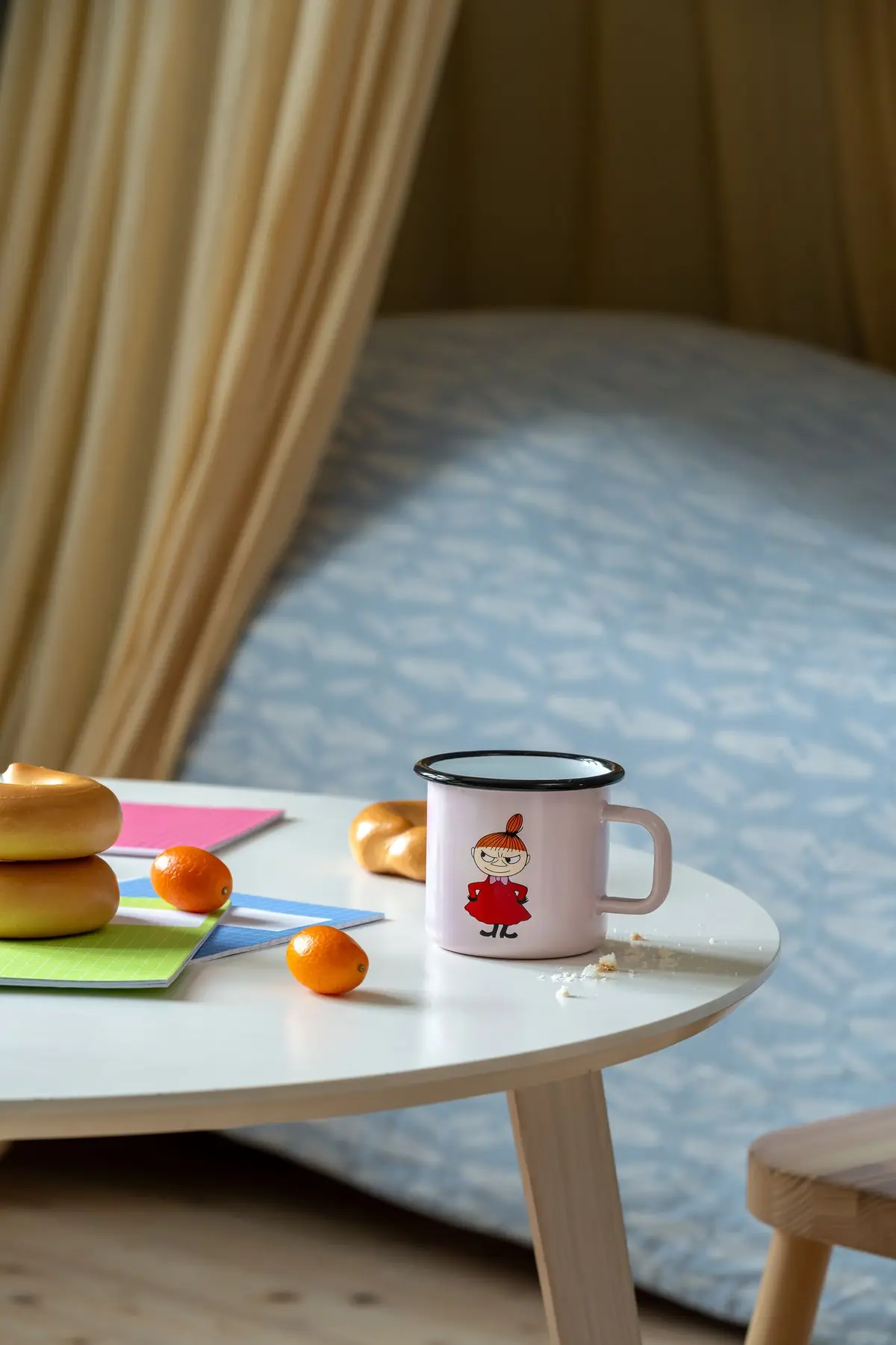 A pink Little MY - Moomin enamel mug (3.7dl) sits on a round table next to bagels, kumquats, and colourful notepads. In the background, there's a bed with a blue patterned blanket. Gate21.ee