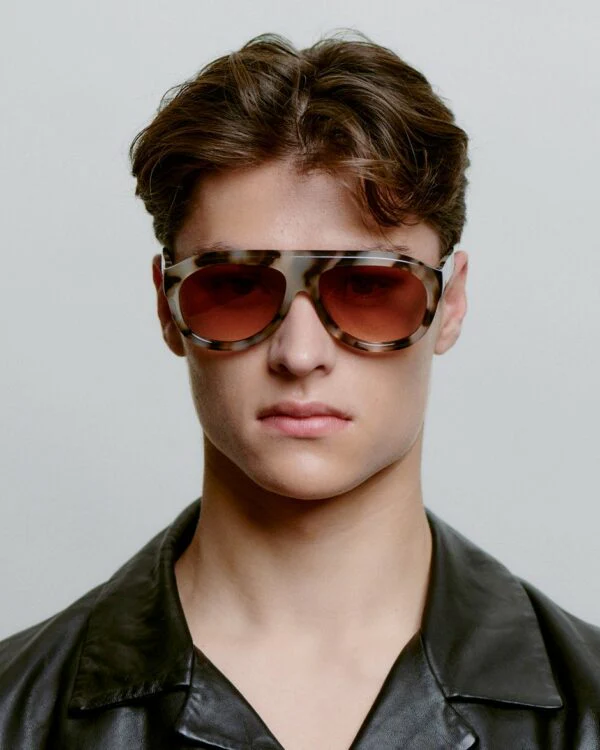 A young person with short, wavy brown hair wears large Rio - Demi Tortoise sunglasses and a black leather jacket, staring directly at the camera against a neutral background. Gate21.ee
