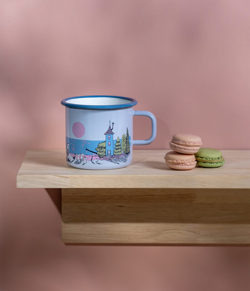 The Sunset - Moomin enamel mug 550ml in light blue with an illustrated scene sits on a wooden shelf beside three stacked macarons—green, pink, and beige—set against a pink background. Gate21.ee