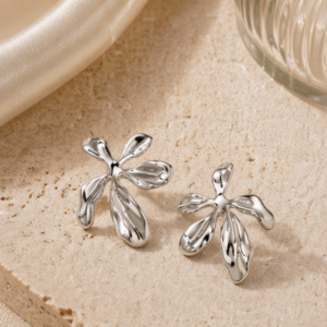 A pair of Nalani - BØLD Earrings in silver, featuring an abstract petal-like design, are displayed on a textured beige stone surface beside a folded cream-coloured fabric and a ribbed glass object. Gate21.ee