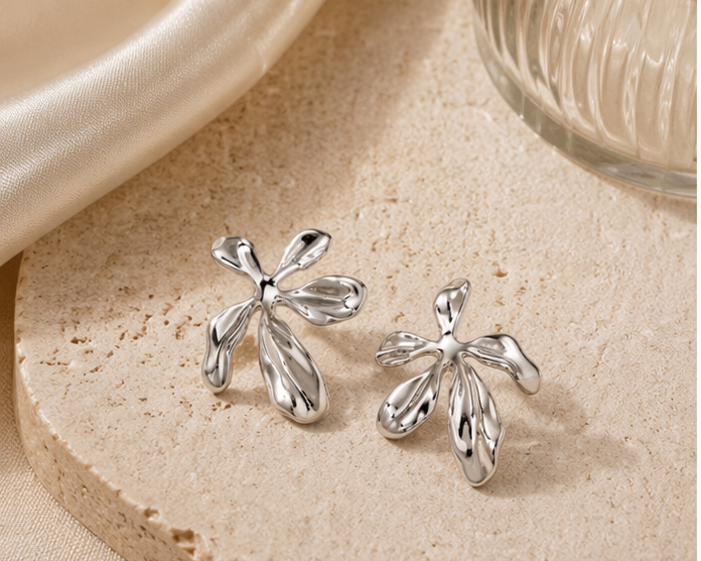 A pair of Nalani - BØLD Earrings in silver, featuring an abstract petal-like design, are displayed on a textured beige stone surface beside a folded cream-coloured fabric and a ribbed glass object. Gate21.ee