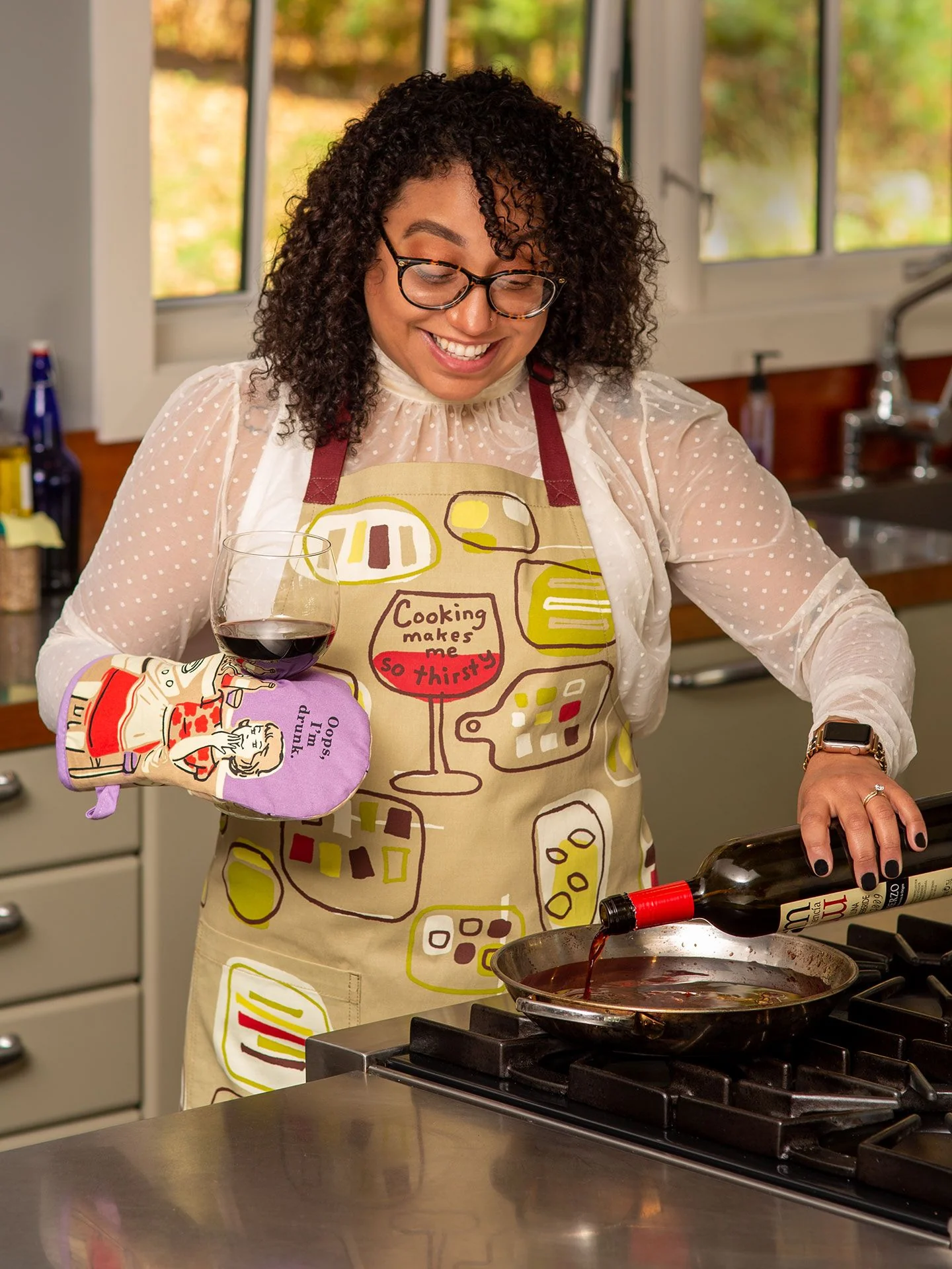 A woman in glasses and an apron smiles in her kitchen as she pours red wine from a bottle into a pan, holding a wine glass and wearing the colourful "Oops, I'm drunk" Oven Mitt by BlueQ. Gate21.ee