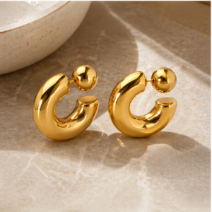 A pair of Dual Gravity - BØLD Earrings in gold, featuring thick, rounded hoops with small spherical studs, displayed on a light stone surface near a beige dish. Gate21.ee