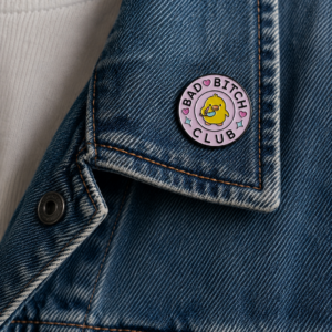 The Bad Bitch Club - Pin, a round enamel badge with a yellow smiley face, is attached to the collar of a blue denim jacket over a white ribbed vest. Gate21.ee