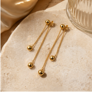 The Dual Gravity - BØLD Earrings - Gold, featuring long chains with small gold spheres at each end, are displayed on a beige stone surface beside a glass and satin fabric. Gate21.ee