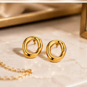 A pair of Halo - BØLD Earrings - Gold, featuring irregular shapes, rests on a marble surface beside a gold chain necklace and a reflective glass object with a gold frame. Gate21.ee