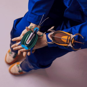 Person in blue clothing sits with hands resting on lap, wearing vibrant StudioROOF Elephant Beetle models in green, blue, brown, and orange on their hands and arm. Shoes are beige with orange and green accents. Background is light-coloured. Gate21.ee
