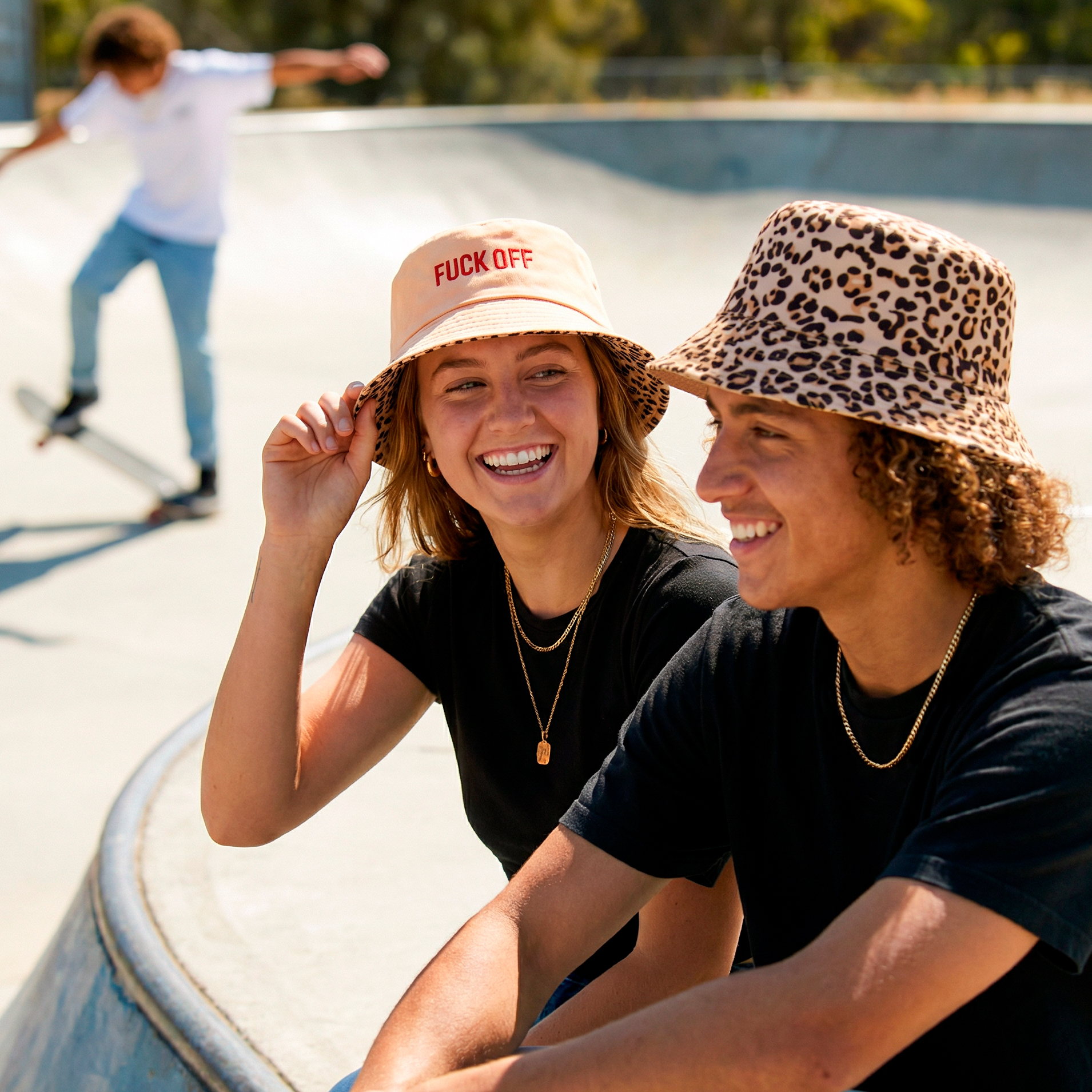 Two young people in bucket hats sit at a skatepark, smiling—one wears the Reversible Bucket Hat - "Fuck Off" with red letters, while another's is leopard print. A handheld fan cools them down as someone skateboards in the background. Gate21.ee