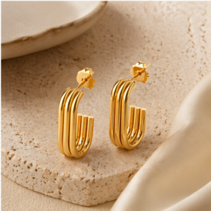 A pair of Dual Gravity - BØLD Earrings in gold, featuring a triple-bar design and a shiny gold-tone finish, rest on a textured beige stone surface beside satin fabric and a ceramic dish. Gate21.ee