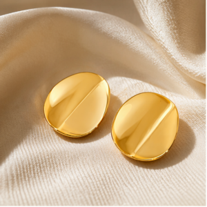 A pair of Luna Plate - BØLD Earrings in gold rests on a smooth, light beige fabric with a subtle sheen. These shiny, oval-shaped earrings feature a simple, modern design accented by a central raised line. Gate21.ee