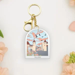 A Võtmehoidja – Talvine mets keyring is placed on a white surface, surrounded by peach flowers, a New Rebels small rucksack, and scattered pastel heart shapes. Gate21.ee