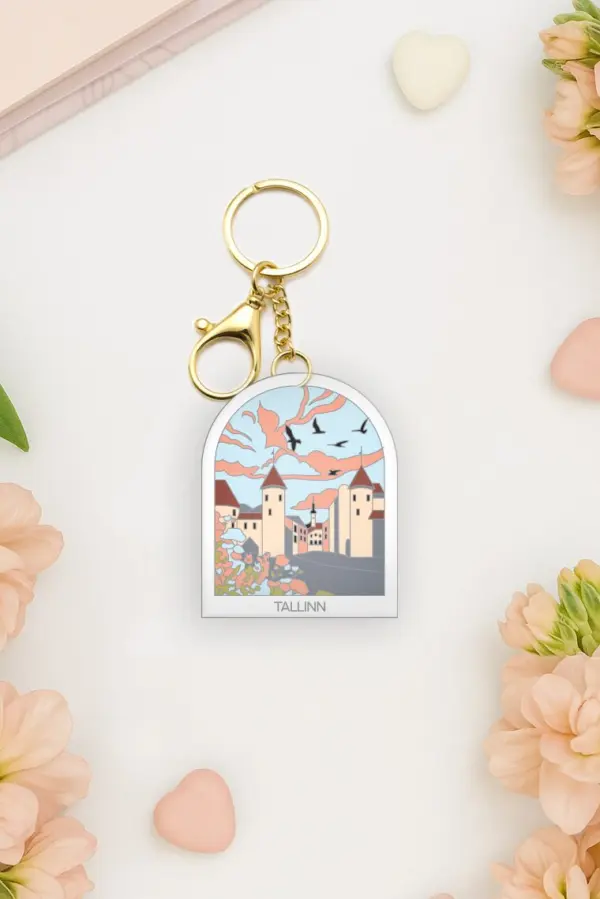 A Võtmehoidja – Talvine mets keyring is placed on a white surface, surrounded by peach flowers, a New Rebels small rucksack, and scattered pastel heart shapes. Gate21.ee