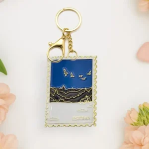 The Võtmehoidja – Talvine mets is a gold keyring shaped like a postage stamp, featuring a blue sky, birds, mountains, the word “Estonia,” and surrounded by pink flowers and heart-shaped objects on a light surface—making it a charming accessory to pair with your New Rebels small rucksack. Gate21.ee